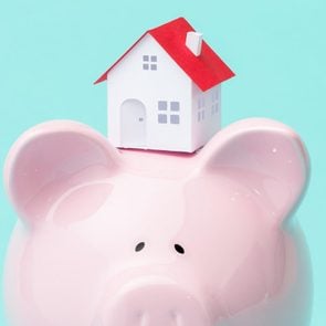 small house sitting atop a piggy bank against teal background