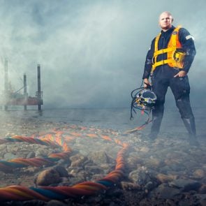 Chris Lemons standing in deep sea diving gear in front of fog that shrouds background. Cords on the ground in the foreground