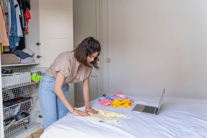Young woman with laptop placing clothes on bed