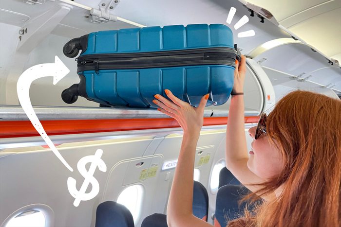 Close-up image of redheaded woman wearing sunglasses, placing wheelie suitcase in airplane cabin overhead baggage compartment, row of seats, focus on foreground