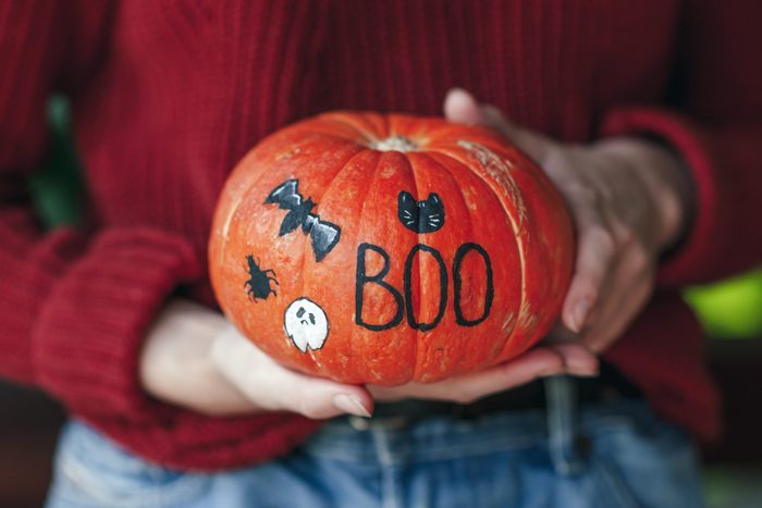 Woman holding a pumpkin painted for halloween; pumpkin is painted with the letters "BOO", a bat, spider, cat, and ghost