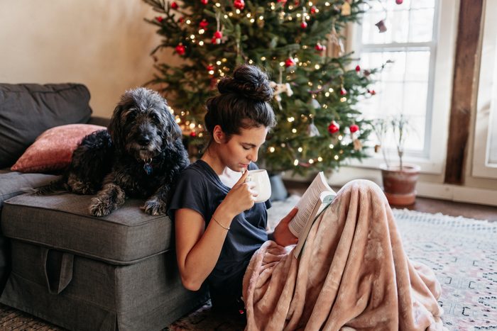 young woman reading and drinking tea with dog by Christmas tree