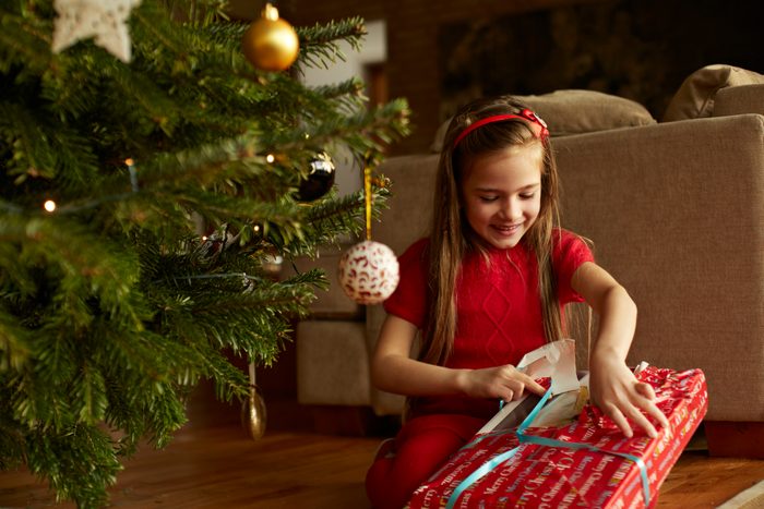young girl opening christmas present next to christmas tree at home