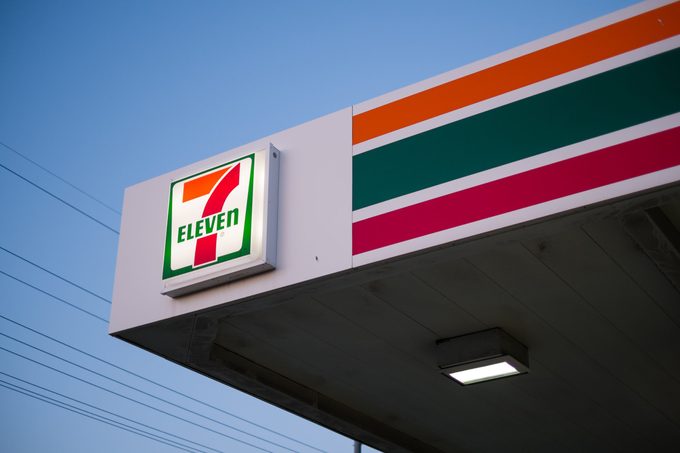 A sign for the popular convenience store chain 7 Eleven on the corner of the awning above gas pumps appears with a clear blue sky and power lines in the background.