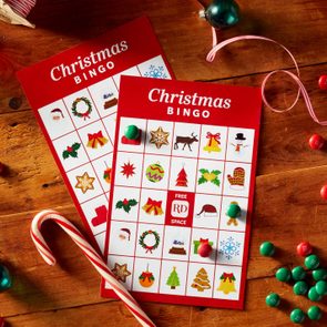 two RD printable christmas bingo cards on a wooden background surrounded by christmas items; candy cane, red and green m&ms, ribbon, christmas ornaments, pinecones