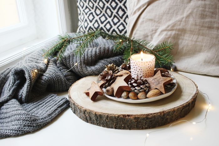 Winter festive still life scene. Burning candle decorated by wooden stars, hazelnuts and pine cones standing near window on wooden cut board. Glittering Christmas lights. Fir branch on wool plaid.