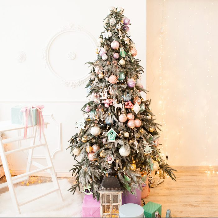Christmas tree decorated with glass and wooden vintage toys and gift boxes in pastel colors near window in light interior with lights garland on wall