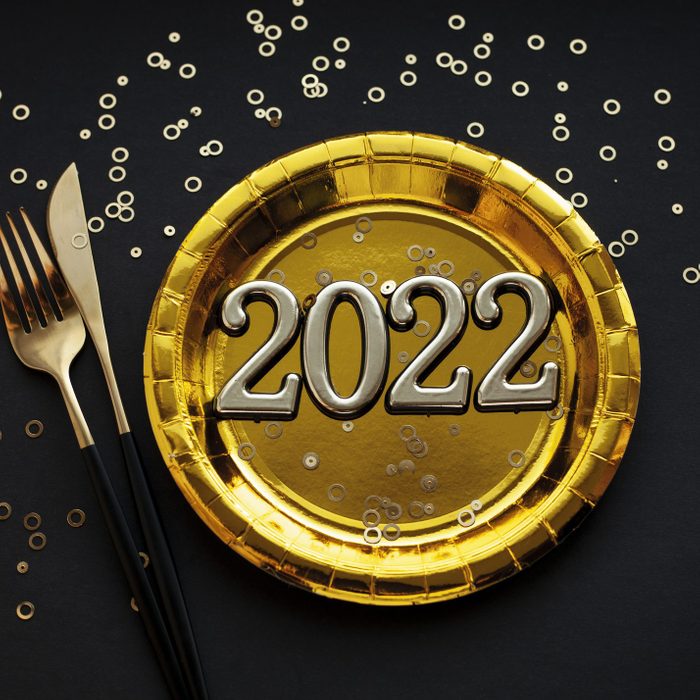 New Year 2022 Greeting Card. Number 2022, golden plate, fork and knife over black background with confetti. New Year party concept.