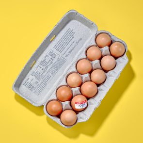 open carton of eggs on yellow background. one egg has a shelf life sticker that has three question marks written on it