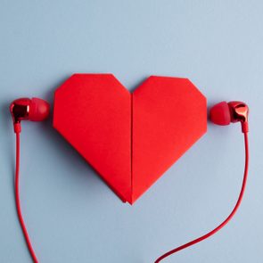 Red origami heart with red earbud headphones on blue background. Listening to valentine's day playlist.