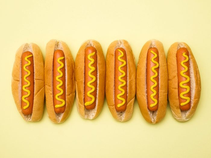 six Hot dogs in buns with mustard against a yellow background