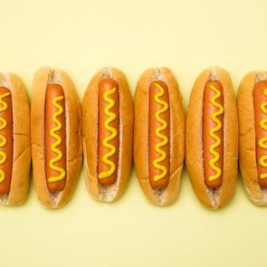 six Hot dogs in buns with mustard against a yellow background