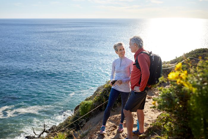 Full length smiling senior couple talking while standing on cliff by sea during sunny day