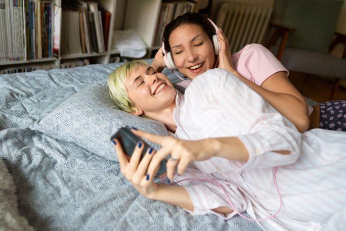 Smiling lesbian couple lying in bed together and listening to the music on a smartphone