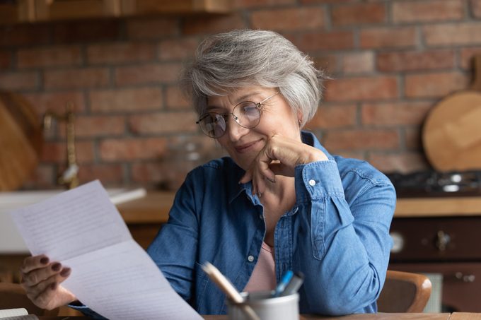 Mujer mayor sentada en la mesa de la cocina leyendo un documento en papel