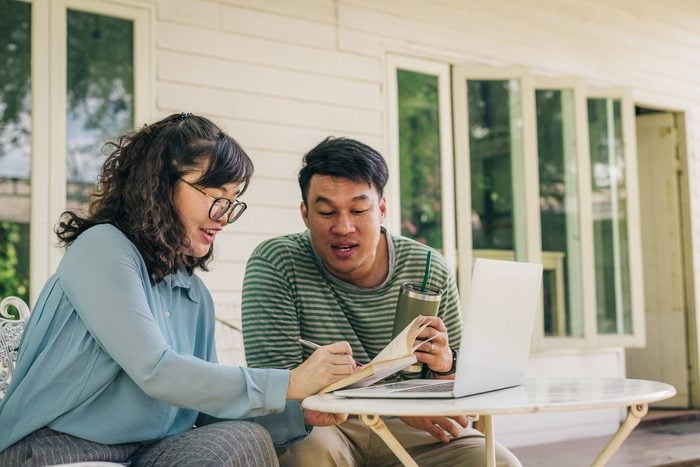 Happy young Asian couple using laptop computer and writing on notebook while sitting in front of the house, Couple relaxing and spending time together in backyard at home, People and lifestyle concept