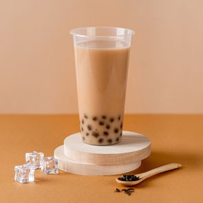 studio shot of milk bubble tea with tapioca in a plastic cup on wood props with prop ice and a small wooden spoon nearby; tan background