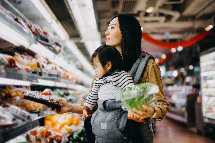 Young mother grocery shopping with baby girl in supermarket, shopping for fresh organic vegetables and fruits