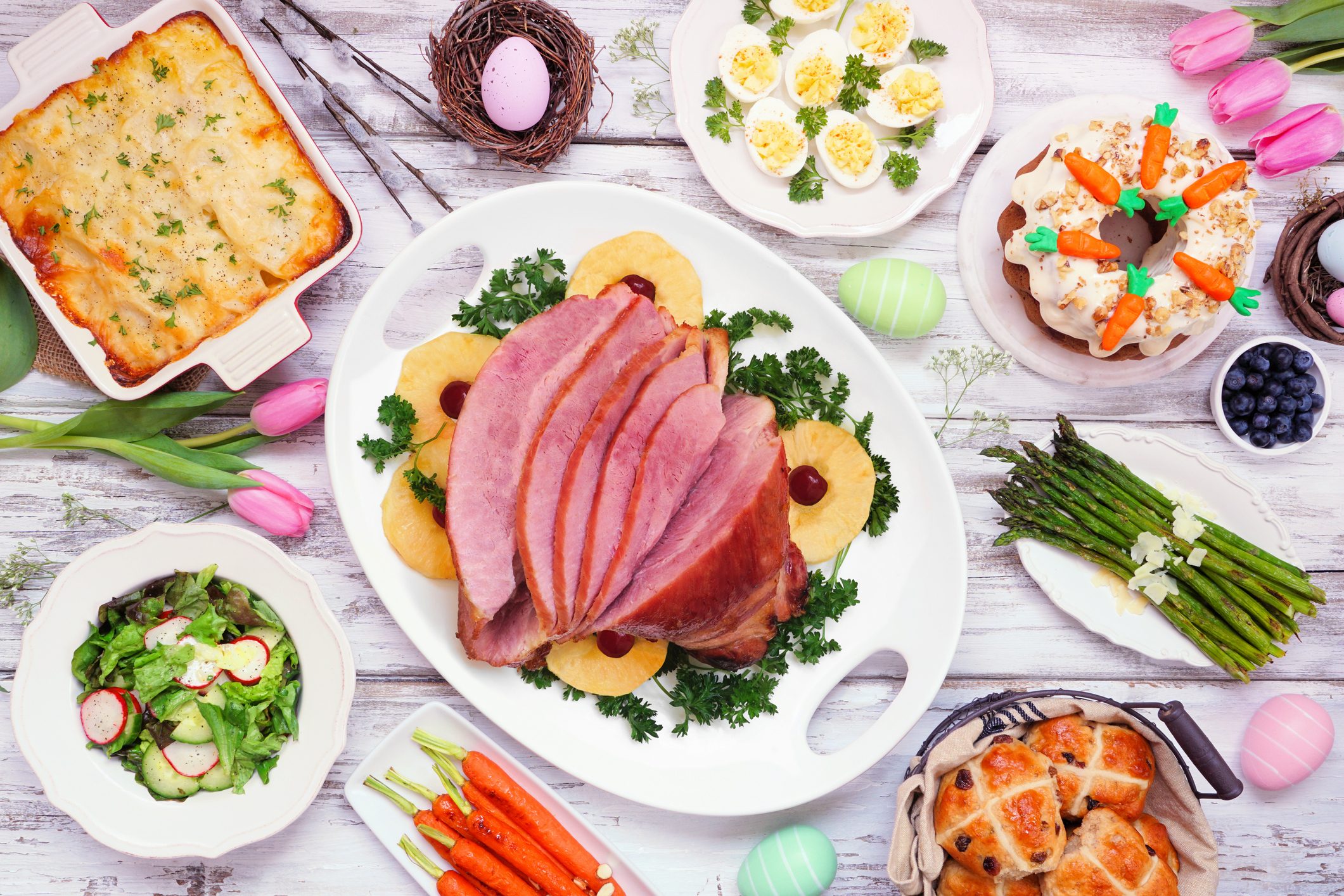 TraditionaTraditional Easter ham dinner. Top view table scene on a white wood background. Ham, scalloped potatoes, eggs, hot cross buns, carrot cake and vegetables.l Easter ham dinner. Top view table scene on a white wood background.