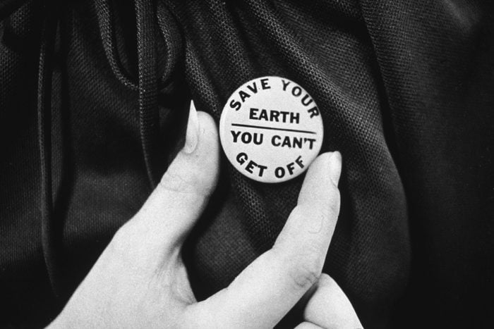 A close-up of a hand holding up an Earth day button which reads, 'Save your Earth - You can't get off.'