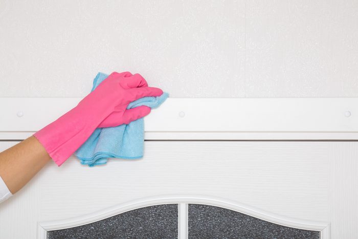 Young adult woman hand in pink rubber protective glove holding blue dry rag and wiping dust from white top frame of wooden door in room