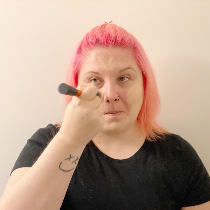 woman with pink hair applying concealer to her face