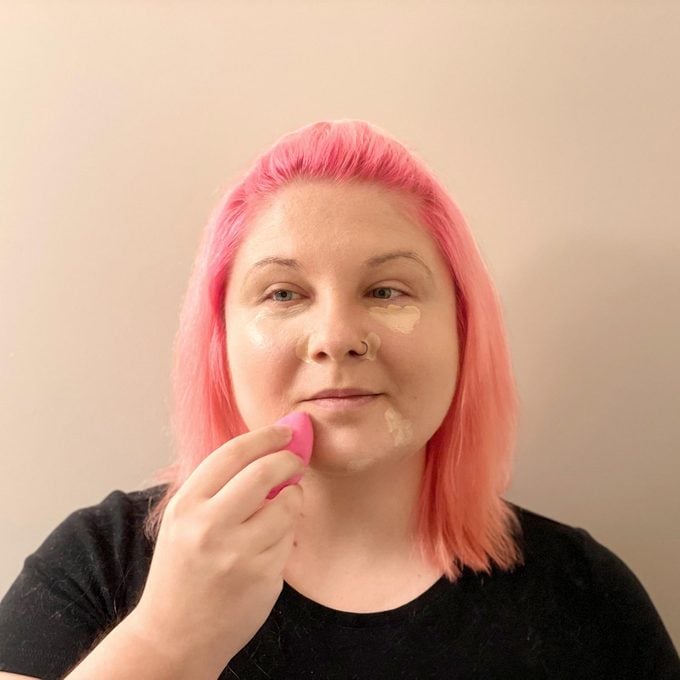 woman with pink hair applying concealer to blemishes on her face