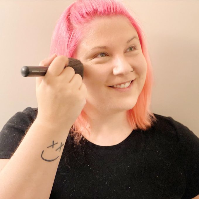 woman with pink hair finishes applying makeup with a a brush and powder
