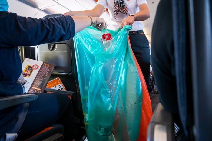 Interior of airplane with passengers throwing plastic bottle into garbage bags held by flight attendant