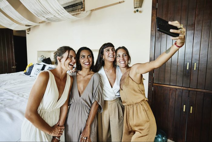 Medium wide shot of smiling and laughing bridesmaids taking selfie in luxury hotel suite before wedding