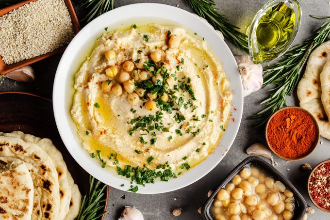 Hummus topped with chickpeas, olive oil and green coriander leaves on stone table with pita bread and spices aside