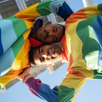 Multi-ethnic gay couple smiling and looking at the camera while hugging, both wearing a hoodie with the LGBT flag.
