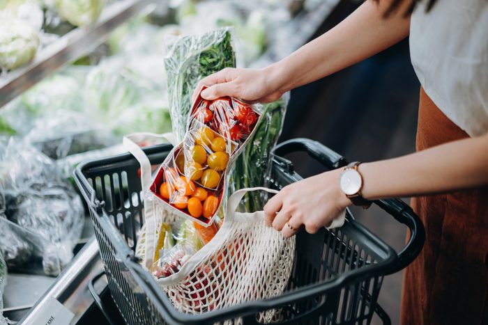 Cropped shot of young Asian woman shopping for fresh organic groceries in supermarket and putting a variety of fruits and vegetables into cotton mesh eco bag in a shopping cart. Zero waste concept
