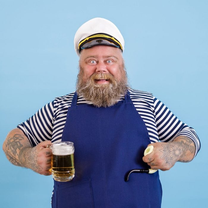 Funny plump person in sailor costume holds smoking pipe and beer on light blue background