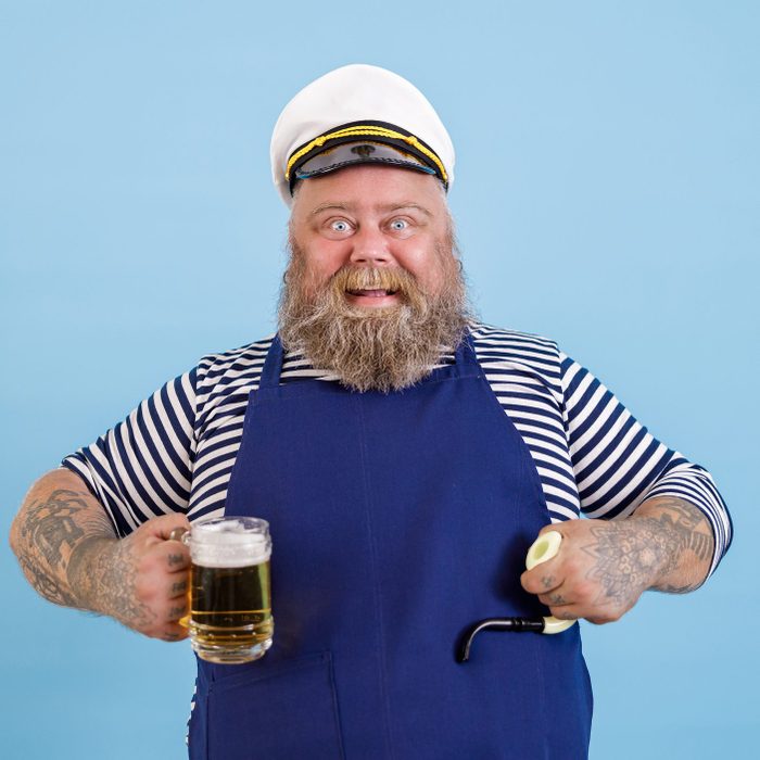 Funny plump person in sailor costume holds smoking pipe and beer on light blue background