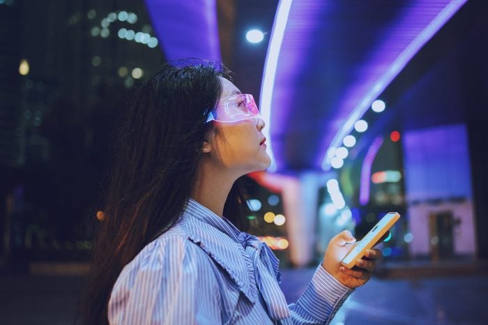 woman using a smartphone and wearing augmented virtual reality glasses while standing under a pedestrian bridge glowing at night