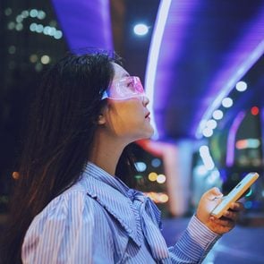 woman using a smartphone and wearing augmented virtual reality glasses while standing under a pedestrian bridge glowing at night