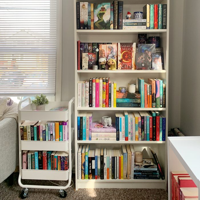 anorganized white bookshelf filled with books and a small metal rolling cart filled with books in front
