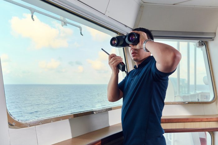 Portrait of cruise navigator pilot officer on the bridge of the vessel looking out the window with binoculars and talking on a radio