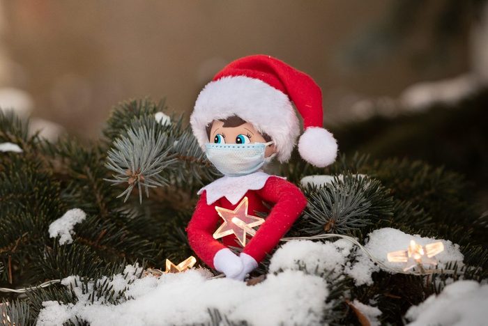 The Christmas toy sits on the branches of a snowy tree in an embrace with a sparkling star. Coronavirus Covid Christmas: a toy doll wearing a real medical mask. Social distancing during a pandemic