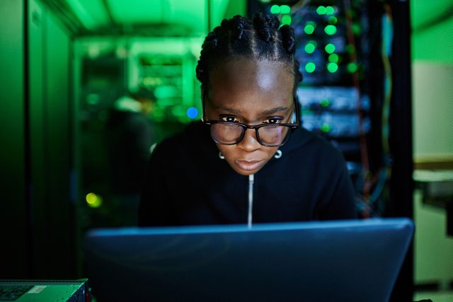 Shot of a young female computer programer using a laptop while standing in a dark server room