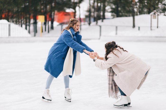 Two happy positive teenage girls friends in warm fur coats are having fun walking skating on a skating rink in the winter forest during the Christmas vacation. A couple of women in love spend time together in nature in cold winter