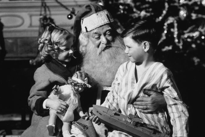 CIRCA 1935: A small boy and girl sit on Santa Claus's lap in front of a Christmas tree. The girl holds a doll and the boy holds a model of a battleship.