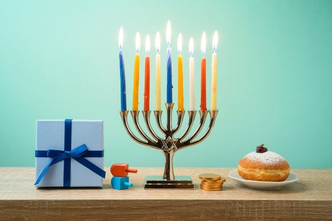 Jewish holiday Hanukkah items including lit menorah, sufganiyot, gift box, dreidels, and gelt on wooden table against light green background