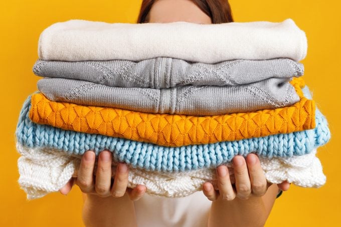 A woman holds a stack of warm knitted clothes on a background of a yellow wall