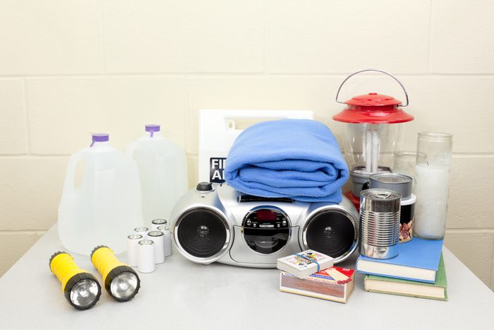 Emergency Kit with flashlights, water, matches, a radio, lantern, candles, books and canned goods on a table with a cement wall in the background