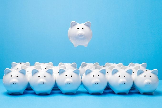 One little white ceramic piggy bank hovering in mid air above group of many other similar piggy banks facing the viewer on blue surface and background