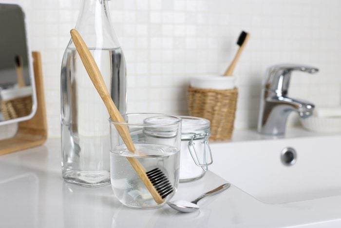 Cleaning a bamboo toothbrush with black bristle in a white vinegar, water and baking soda solution on the bathroom sink close up.