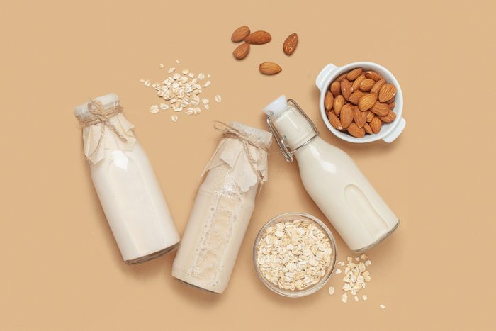 three glass jars of milk with almonds and oats to the side, on a tan background