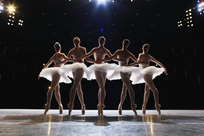 Five ballerinas en pointe on stage, arms around each other, rear view
